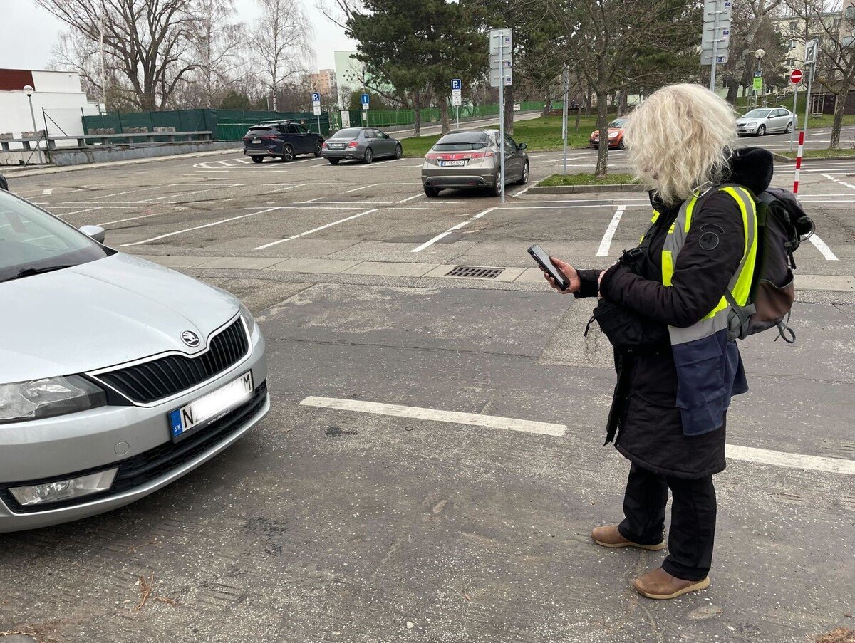 Pani Ivana je parkovacia asistentka. Má na sebe reflexnú vestu a používa zariadenie, ktorým kontroluje zaparkované autá.